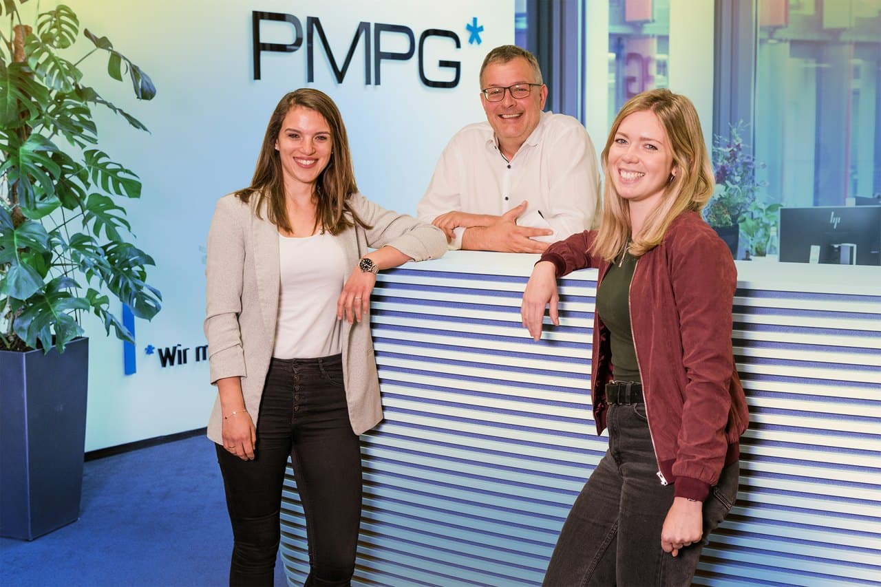Drei PMPG-Mitarbeitende lachend am Empfang vor dem PMPG-Logo